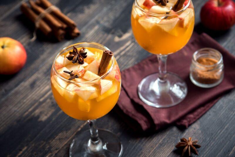 Two wine glasses containing an autumn mead sangria cocktail, garnished with star anise and cinnamon sticks, with more cinnamon sticks and some apples on the table