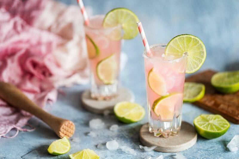 Two tall glasses with pink lemonade vodka, ice, and lime slices, on a light blue table