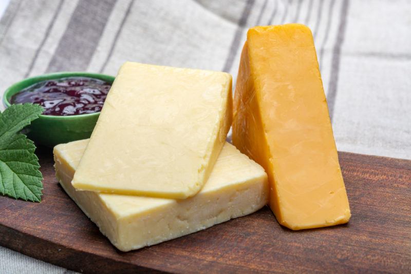 Two types of aged cheddar on a wooden board next to jam or another type of spread