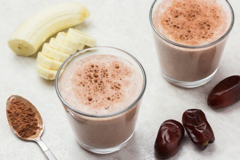 A white plate with two chocolate protein smoothies, next to some bananas and dates