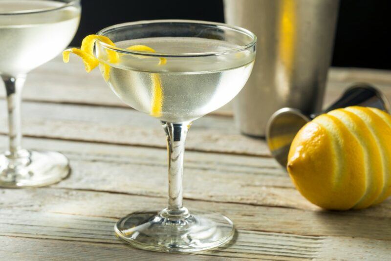 Two vesper martinis on a light wooden table, with a lemon, a jigger, and a cocktail shaker in the background