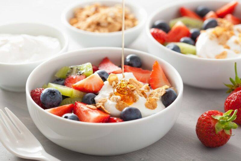 Two large white bowls with breakfast cereal, fruit, and honey, next to a bowl of yogurt and another of cereal