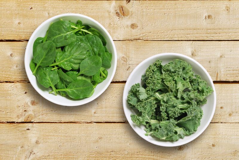 Two white bowls on a wooden table. One contains fresh spinach and the other contains fresh kale.