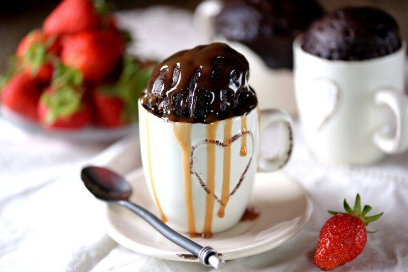 White mugs containing chocolate mug cakes, one with a spoon and caramel sauce dripping down the side