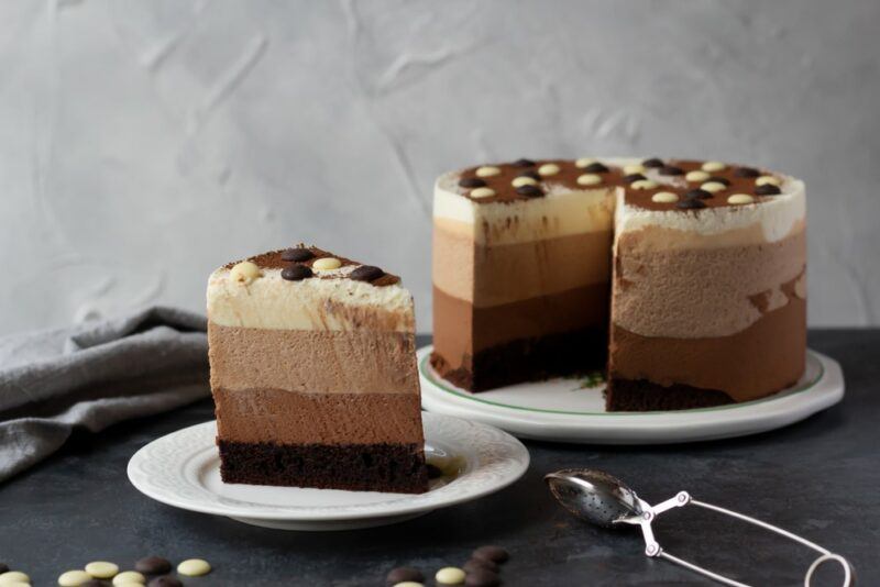 A small white plate with a triple layer chocolate cake, with a large plate containing more cake in the background