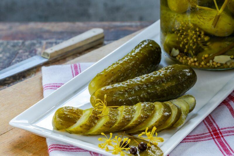 A white rectangular plate with two whole pickles, a pickle that has been sliced into pieces and a jar of pickles in the background