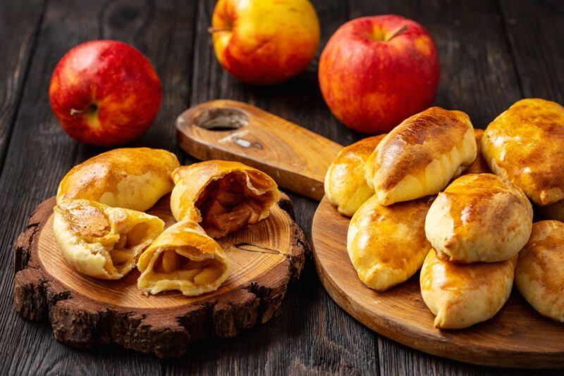 A dark wooden table with three fresh apples, plus a wooden board of sweet pirozhki cakes and some more cakes that have been cut open to reveal sweet fillings