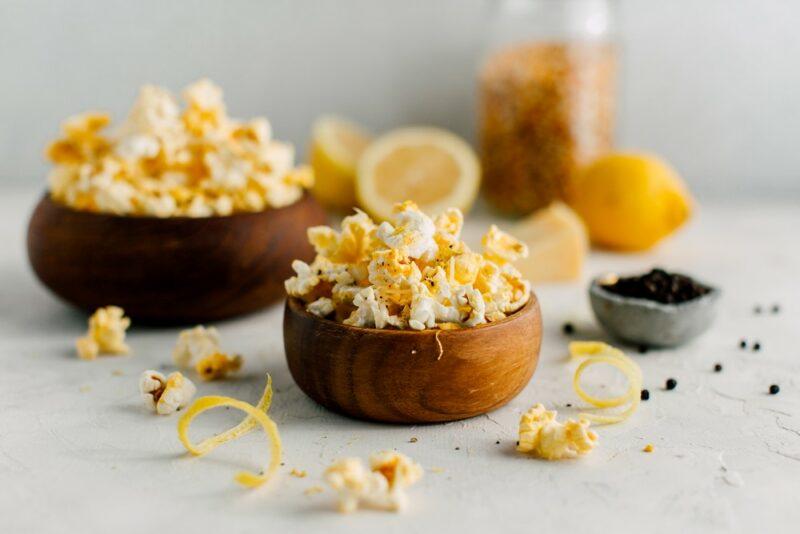 Two wooden bowls containing popcorn with black pepper and lemon, with a bowl of black pepper and some lemons in the background