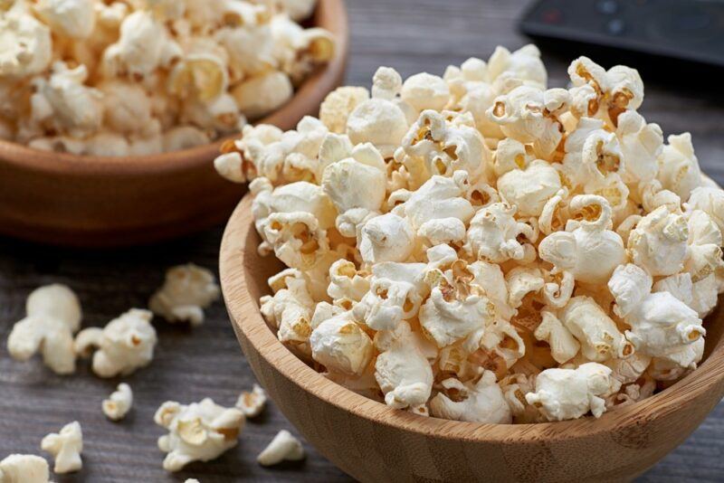 Two wooden bowls filled with plain popcorn, with more popcorn on the table