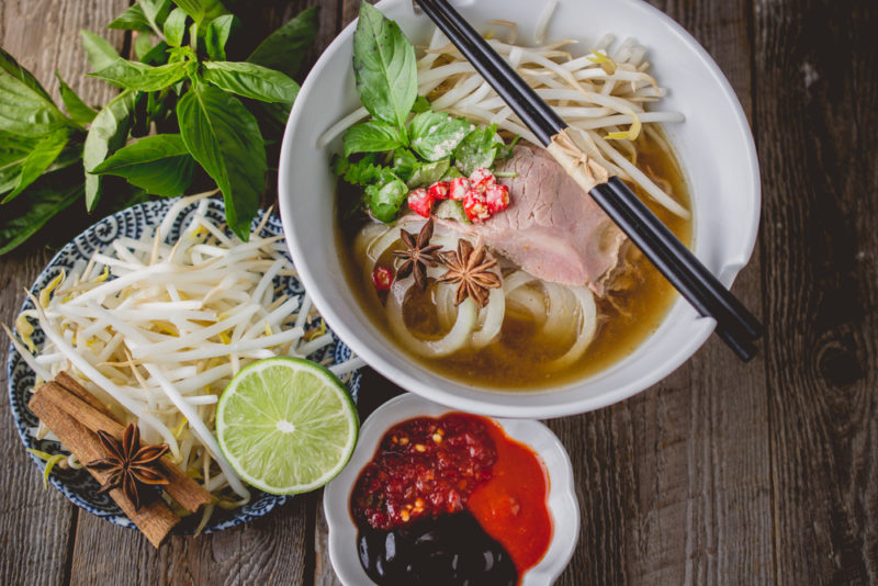 A large white bowl of Vietnamese pho soup next to sauces and other toppings