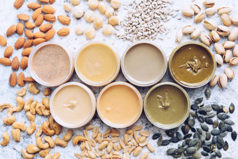 Seven white bowls that each contain a different type of nut or seed butter. Next to each bowl is the appropriate nut or seed