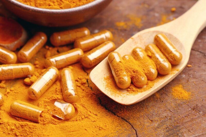 A wooden table with turmeric capsules and powder. Some of the capsules are on a large wooden spoon