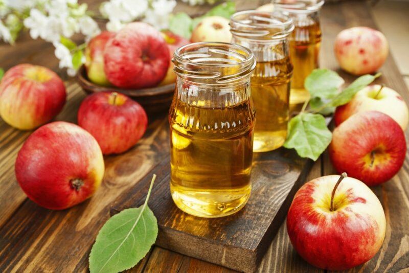 A wooden board with three glasses of apple juice and many fresh red apples