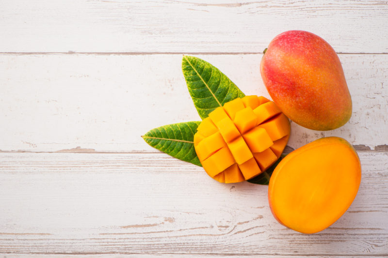 A white table with a whole mango, a half mango, and a mango that has been sliced into chunks