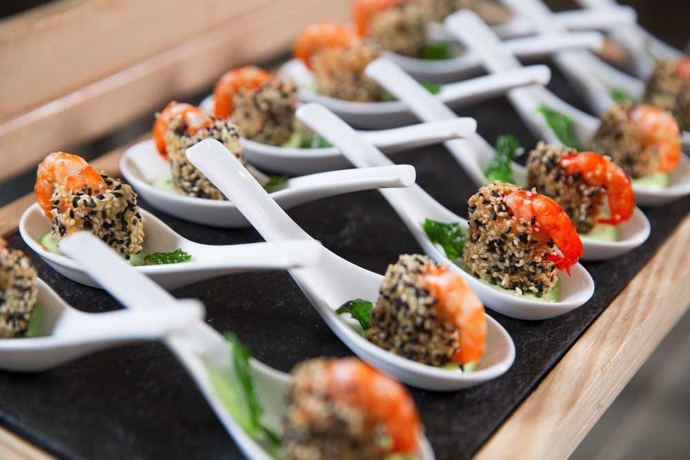 A selection of white spoons on a black plate, where each contain a shrimp appetizer