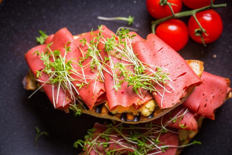 A piece of bread with vegan ham or pastrami and sprouts, with fresh cherry tomatoes in the background