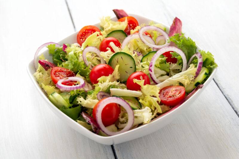 A square white bowl filled with green salad. red cherry tomatoes and purple onion rings rests on a white wooden surface.