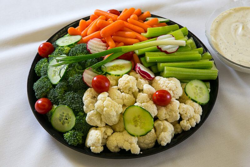 A round tray of veggies, including cauliflower, baby carrots, celery sticks, and broccoli, rests on a white cloth near a dish of white salad dressing dip.