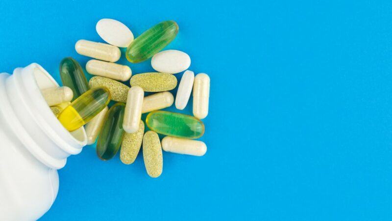 A selection of multivitamins spilling out of a white bottle against a bright blue background