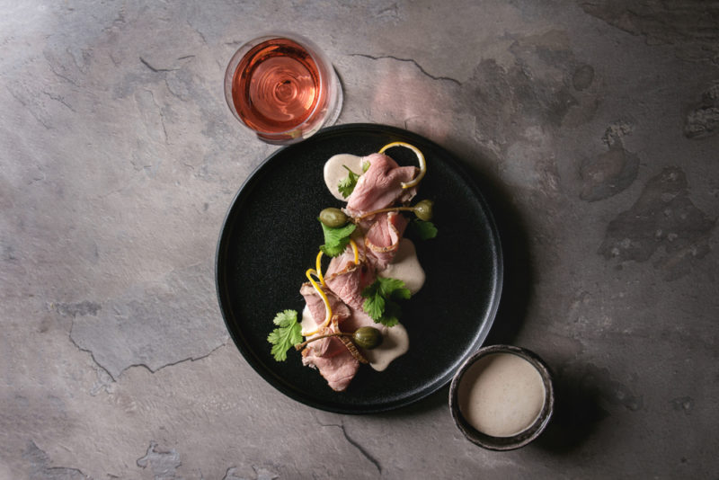 A black dish with vitello tonnato next to a glass of wine and some dressing