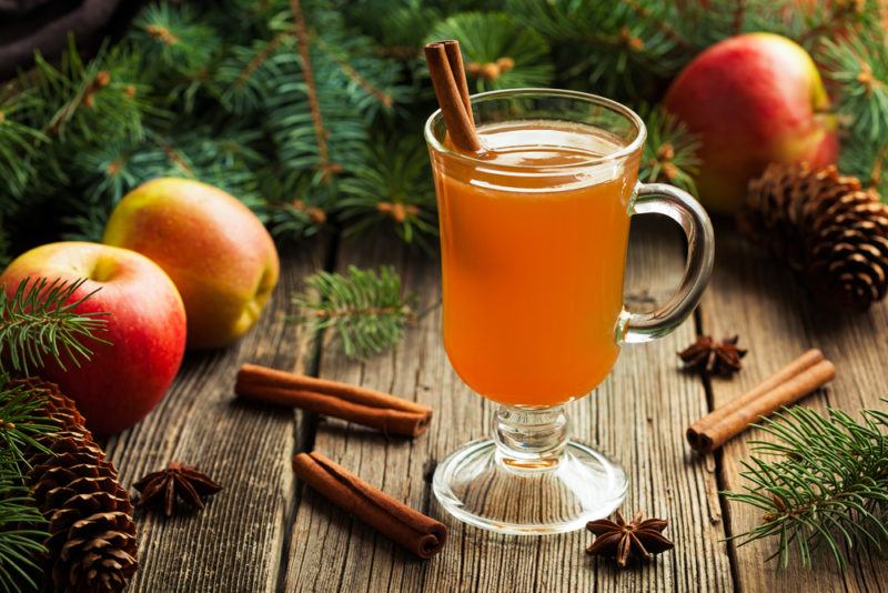 A glass mug of warm cider next to some apples and cinnamon sticks, in front of pine leaves