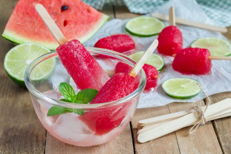 A glass bowl of watermelon ice blocks on sticks, with more ice blocks and some watermelon and limes in the background