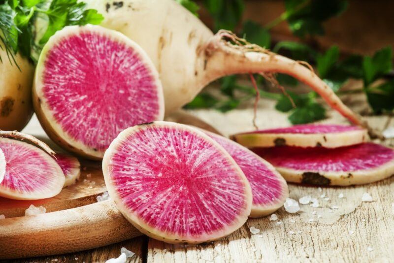 A wooden table and board containing a whole watermelon radish and some slices of the vegetable