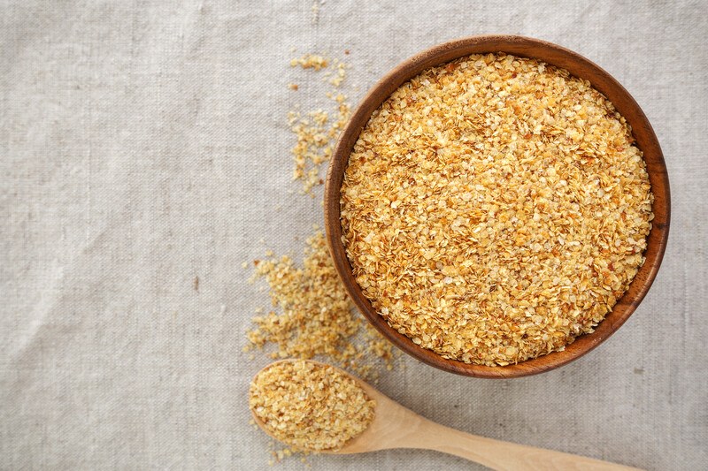 This overhead photo shows a wooden bowl of wheat germ and an overflowing wooden spoon of wheat germ on a beige cloth.