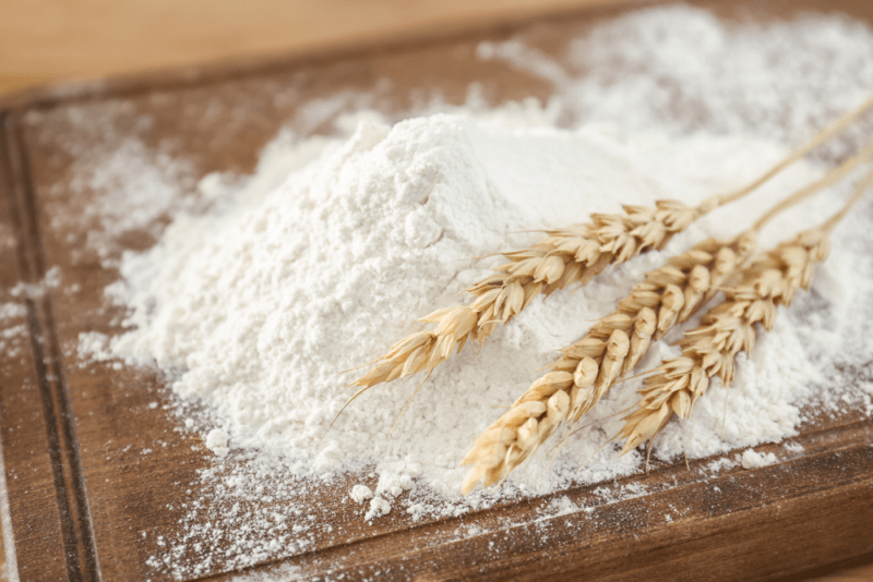 A wooden table with wheat flour, plus a few stalks of wheat.