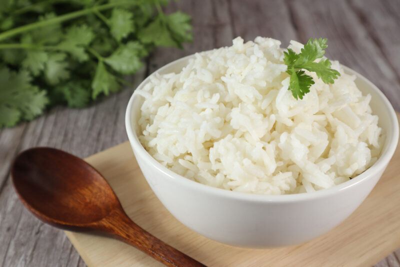 A bowl of cooked white rice with a sprig of parsley rests on a cutting board near a wooden spoon and more parsley on a wooden table.