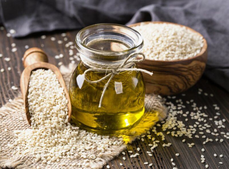 A small glass container of sesame seed oil, next to a bowl and a scoop of the seeds