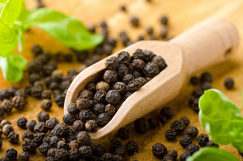 Closeup image of whole peppercorns in a wooden scoop resting on a wooden table.