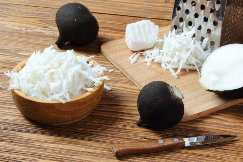 A wooden table with some whole black radishes, one that has been cut in half and some grated radish