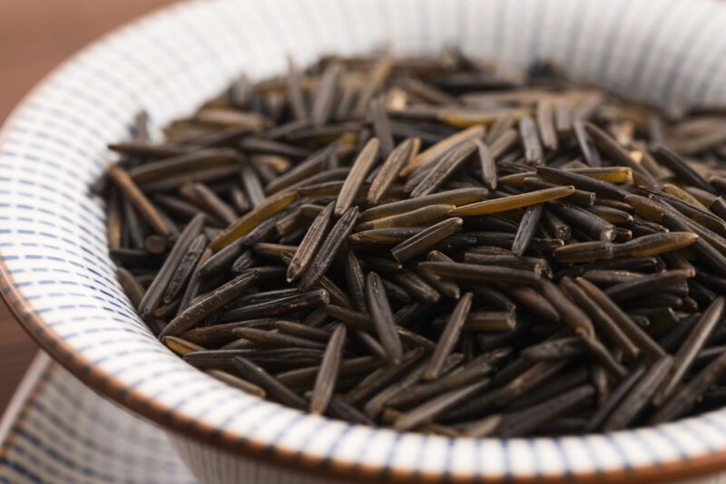 on a wooden surface is a closeup image of a white ceramic bowl with brown rim full of wild rice