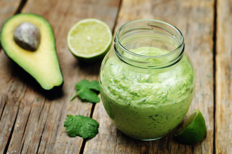 A glass jar of avocado dressing next to half an avocado and half a lime