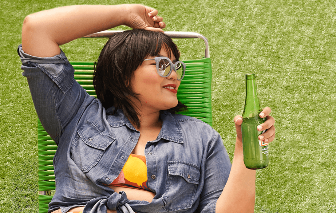 Woman smiling looking off to right while sitting on a outdoor lounge chair holding a bottle of soda, wearing a denim top tied in the front with green turf in the background