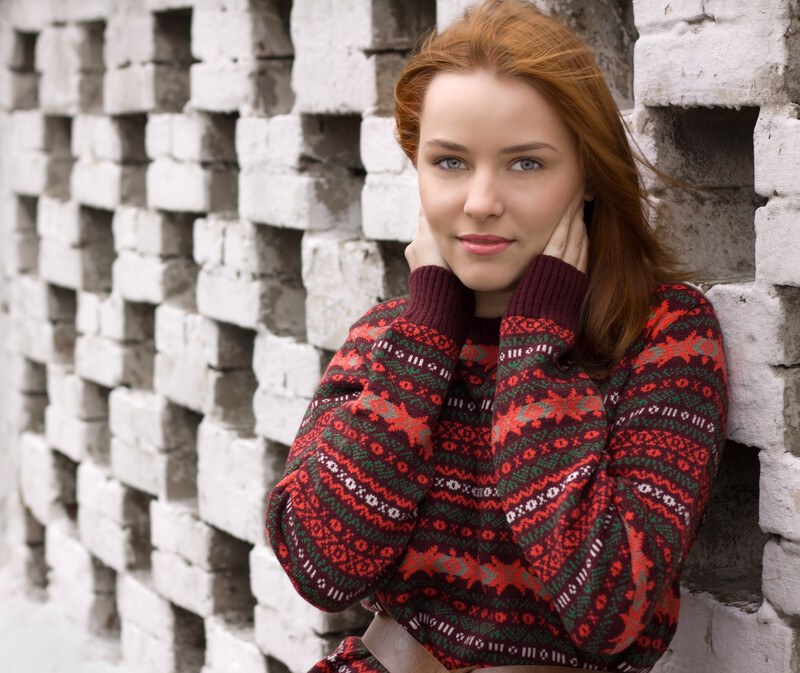 Woman with red hair smiling at the camera wearing a colorful sweater leaning against a brick wall