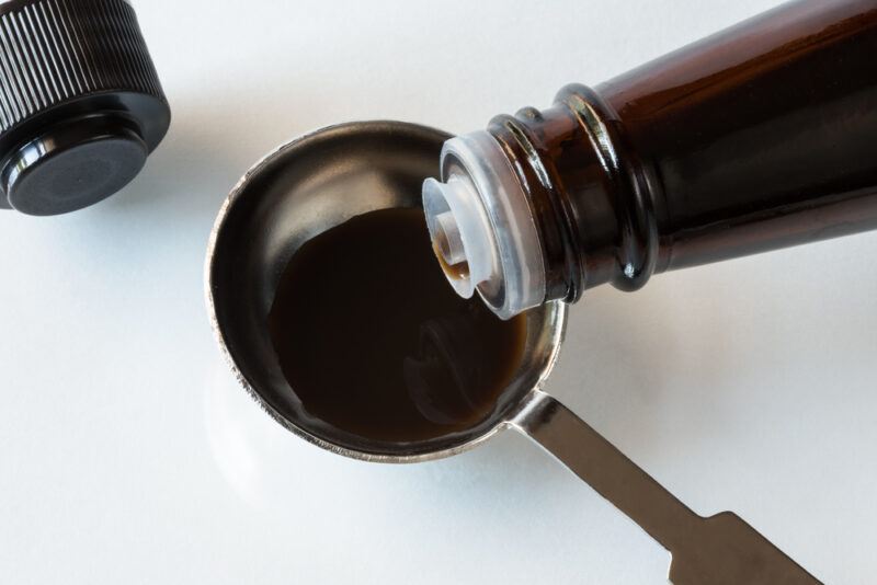 overhead shot of white surface with a bottle of Worcestershire sauce with its content being poured in a metal measuring spoon, beside it is a black bottle cap