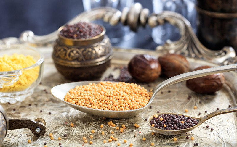 On a sliver tray with scattered mustard seeds are white-yellow mustard seeds in glass bowl, brown, and black mustard seeds in silver spoons, and a little jar of black mustard seeds.