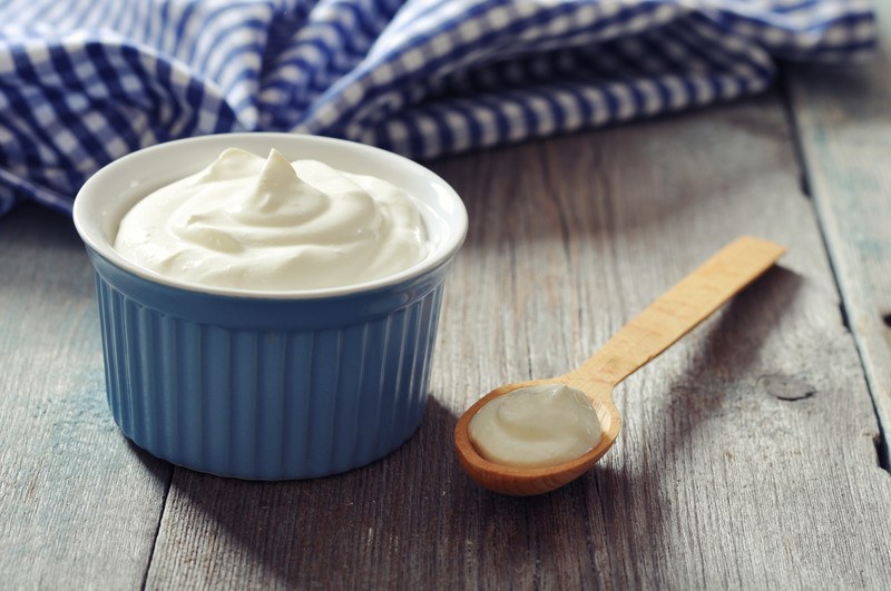 This photo shows a blue ceramic bowl filled with Greek yogurt on a wooden table next to a blue and white cloth and a wooden spoon with yogurt in it.