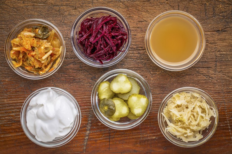 This photo shows an overhead view of six open jars of fermented foods, including kimchi, beets, apple cider vinegar, yogurt, and sauerkraut.