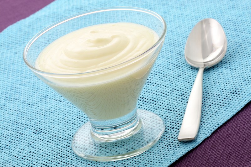 A clear dish of white yogurt rests on a blue cloth next to a silver spoon.