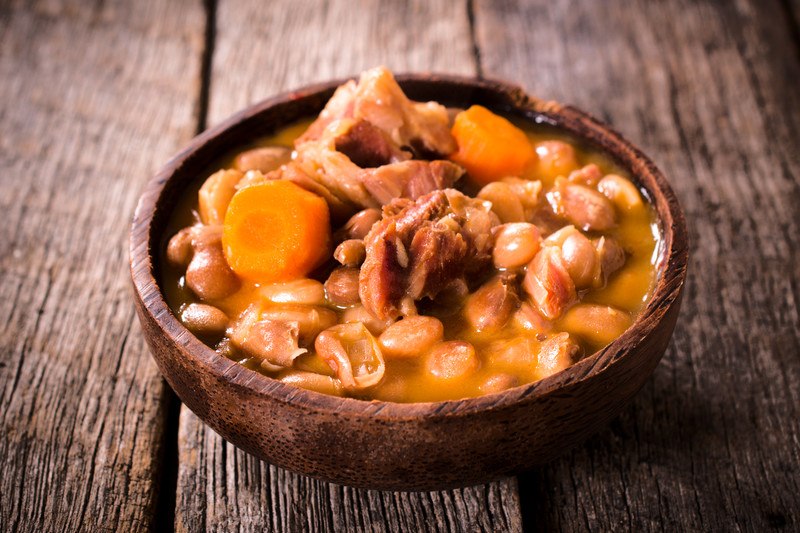 This photo shows a wooden bowl of beef, beans, and carrots sitting on a wooden tabletop.