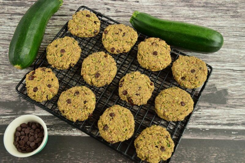 A wire baking tray with 12 zucchini Martian cookies cooling, next to a small bowl and two zucchinis