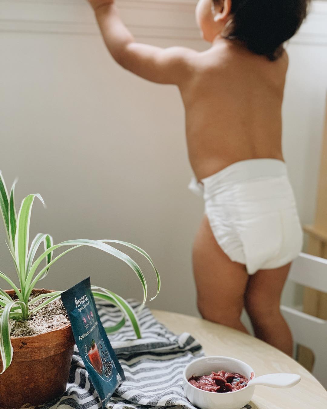 a baby standing holding onto the window looking up and out with a small table in front with a white bowl and spoon full of baby food