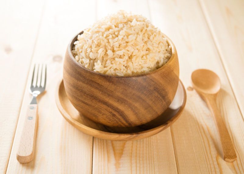 on a wooden natural-colored surface, is a wooden bowl of brown rice, resting on a wooden plate with wooden spoon and fork with wooden handle