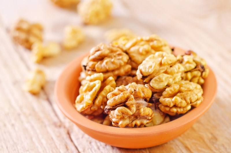 An orange dish full of shelled walnuts sits on a wooden table next to a handful of loose shelled walnuts.