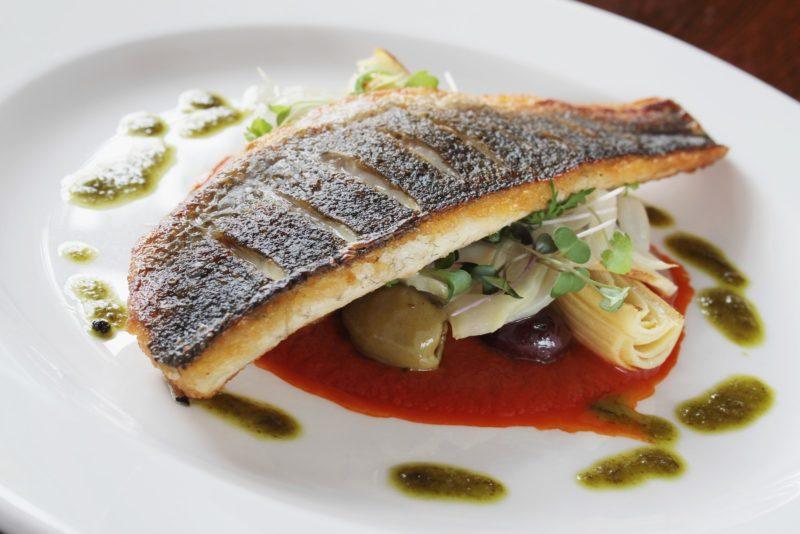 grilled seabass fillet served on a white plate with some olives, onions and two different types of sauce