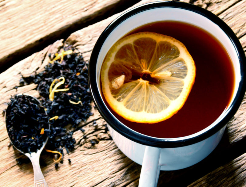 A white ceramic mug of black tea with a slice of lemon, next to some tea leaves