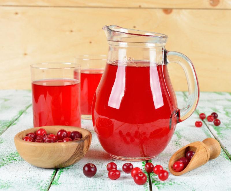 one pitcher of cranberry juice, two glasses filled with cranberry juice, fresh cranberries on a bowl, all placed on a wooden table